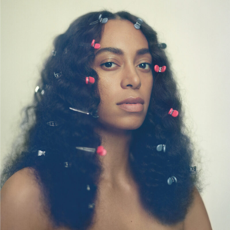 Retrato aproximado de Solange, de frente para a câmera. Ela tem os cabelos soltos e volumosos, presos por diversos grampos coloridos, principalmente rosas e prateados. Usa maquiagem discreta e expressão serena. O fundo é claro e desfocado.
