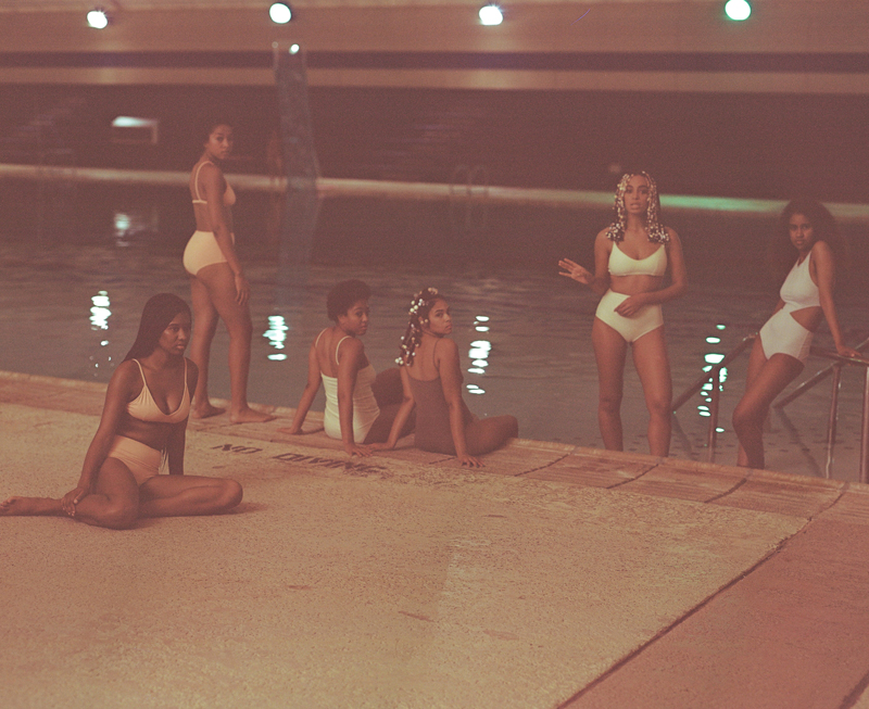 Foto de uma piscina em ambiente interno, com cinco mulheres negras usando biquínis claros. Algumas estão sentadas na borda da piscina, enquanto outras permanecem em pé dentro ou próximas à água. A mulher ao centro usa biquíni verde-claro e tem o cabelo decorado com diversas contas. A imagem tem iluminação baixa e uma tonalidade rosada, com reflexos das luzes na água.