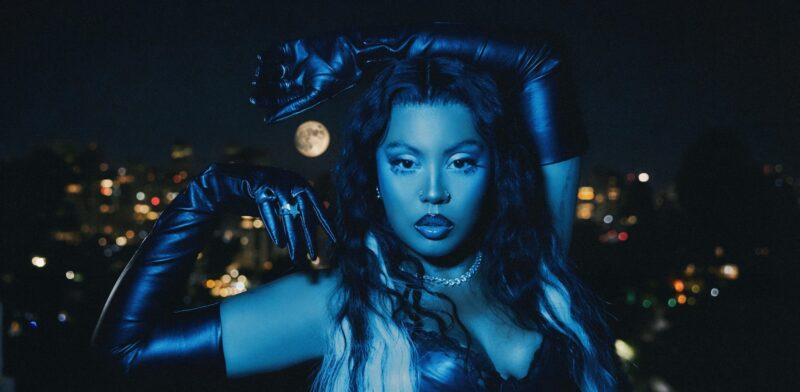 Budah posa diante de uma paisagem urbana noturna iluminada por pontos de luz desfocados. A artista é iluminada por uma luz azul intensa e encara diretamente a câmera com expressão séria. Veste luvas pretas longas, colar prateado e um figurino escuro, enquanto seus cabelos longos apresentam mechas claras na parte frontal. Com os braços erguidos, ela posiciona as mãos de forma a enquadrar visualmente a lua cheia ao fundo, criando a ilusão de segurá-la entre os gestos. A fotografia integra a identidade visual do álbum Frequência Lunar, explorando elementos simbólicos ligados à noite, à lua e à atmosfera mística que caracteriza o projeto