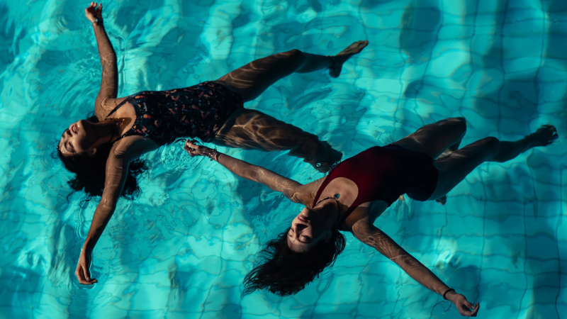Cena de Chicas Tristes. Plano zenital que mostra duas mulheres boiando de costas sobre a água azul-turquesa de uma piscina de azulejos. Ambas surgem de braços abertos e olhos fechados com uma expressão tranquila e de entrega. A mulher à esquerda veste uma roupa de banho estampado escuro, enquanto a mulher à direita usa uma roupa de banho liso vermelho. Os reflexos ondulados da água e sombras projetam padrões dinâmicos sobre os seus corpos submersos