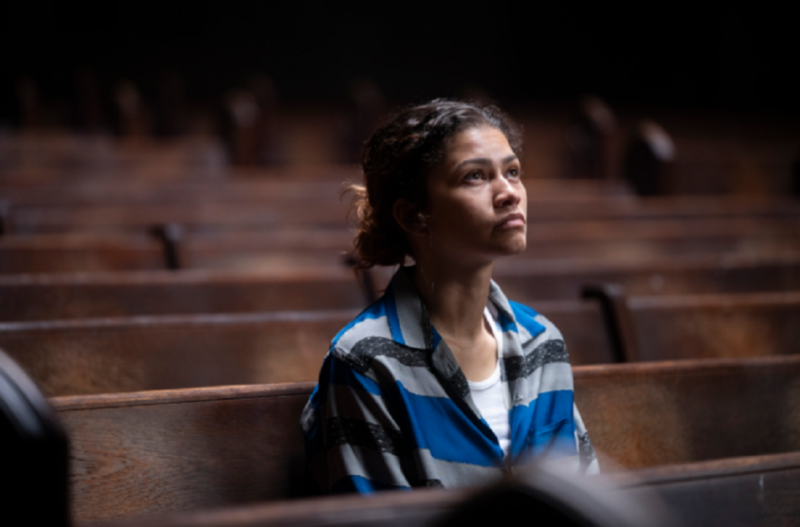Cena da 3° temporada de Euphoria. Plano médio de Rue (Zendaya) sentada sozinha em um banco de madeira escura no interior de uma igreja ou auditório. Ela tem o cabelo apanhado num coque desalinhado e veste uma camisa de mangas compridas com listras largas em azul, cinza e preto, sobre uma camisa interior branca. Rue olha para cima com uma expressão contemplativa, melancólica e atenta. O fundo exibe várias filas de bancos vazios sob uma iluminação suave e penumbrosa. 