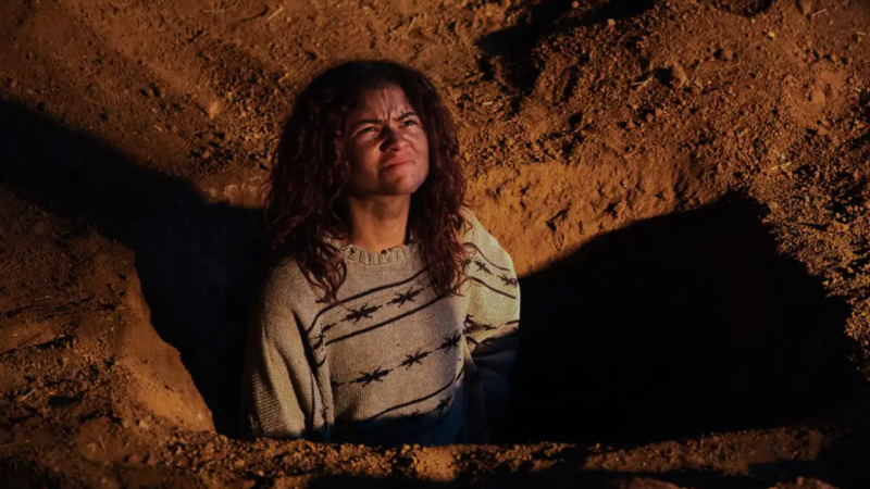 Cena da 3° temporada de Euphoria. Plano médio em ângulo mergulhador mostrando Rue (Zendaya) de pé dentro de uma cova ou vala aberta em terra vermelha e seca. A iluminação lateral intensa realça a textura do solo e projeta uma sombra escura acentuada ao lado dela. Rue veste uma suéter de malha grossa cinza com padrões geométricos e franze o sobrolho enquanto olha para cima com um semblante desgastado e interrogativo. 