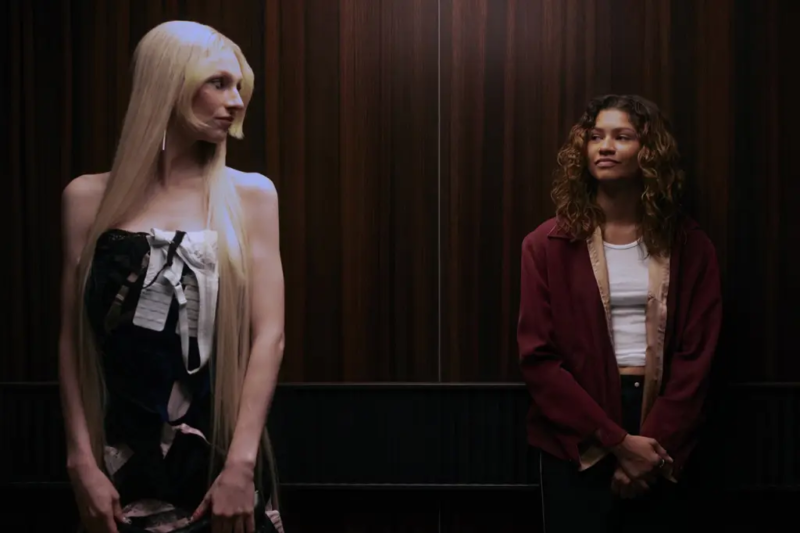 Cena da 3° temporada de Euphoria. Plano médio de Jules (Hunter Schafer), uma mulher branca com cabelo loiro e liso, que chega até a altura da cintura, e Rue (Zendaya), mulher negra, cabelos castanhos e ondulados, que chegam até a altura do ombro. Elas estão lado a lado diante de um painel de parede com ripas de madeira escura. À esquerda, Jules surge com um vestido escuro sem alças e um laço claro no busto, olhando de perfil para Rue. À direita, Rue usa um casaco bordeaux largo sobre uma camisola branca e calças pretas, olhando de volta para Jules com um leve sorriso e as mãos cruzadas à frente do corpo.