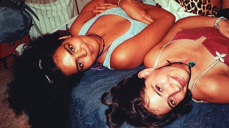 Foto das atrizes de Rocio Guzman e Darana Alvarez. Plano picado aproximado das duas jovens deitadas de costas numa cama com colcha azul escura, com as cabeças voltadas para a câmara e cabelos escuros espalhados. À esquerda, Rocio Guzman, uma jovem de pele morena e cabelos encaracolados veste um top azul-claro brilhante, encarando a lente com uma expressão serena. À direita, Darana Alvarez, outra jovem, surge com a cabeça deitada para o lado e sorrindo abertamente, vestindo um corpete vermelho com alças de pérolas decoradas com um laço prateado e um colar com pendente de coração azul. A fotografia tem uma iluminação direta de flash e textura nostálgica de película analógica.