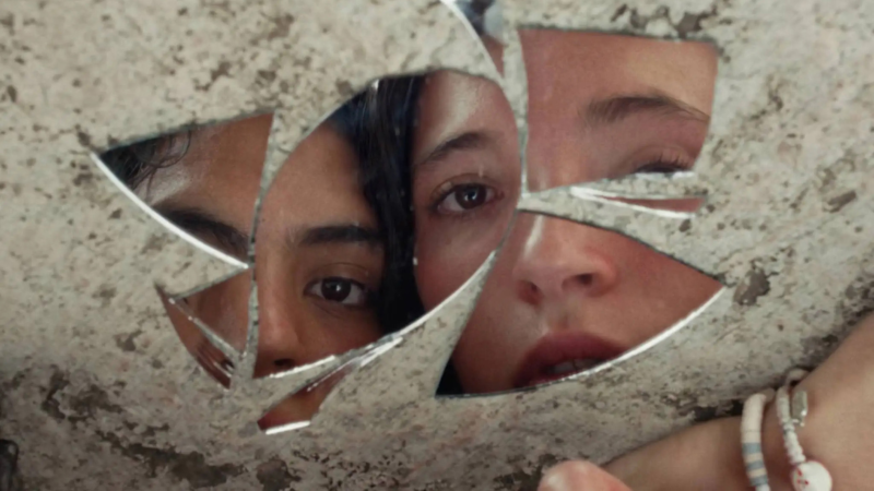 Cena de Chicas Tristes. Grande plano detalhado de fragmentos pontiagudos de um espelho partido sobre uma superfície de pedra clara. Nos cacos de vidro refletem-se, de forma fragmentada, os rostos de Rocio Guzman e Darana Alvarez: no pedaço da esquerda vê-se um olho escuro atento de Rocio Guzman, enquanto nos pedaços centrais e da direita surgem fragmentos de nariz, lábios entreabertos e olhar compenetrado de Darana Alvarez. No canto inferior direito, vê-se parte de um pulso com pulseiras de miçangas e pequenos amuletos.