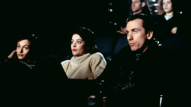 Cena de Fuga para Odessa. Plano médio de três pessoas sentadas lado a lado numa sala de cinema escura, iluminadas pelo reflexo da tela. Em primeiro plano, à direita, Tim Roth tem uma expressão cética e pensativa, vestindo roupa preta. Ao centro, Moira Kelly, mulher branca de cabelo liso escuro, surge com uma camisola de gola alta de lã clara e batom vermelho, olhando atentamente para a frente. À esquerda, Edward Furlong, homem branco de cabelo liso escuro, apoia a cabeça na mão com ar distraído, enquanto outros espectadores aparecem desfocados nas filas de trás.