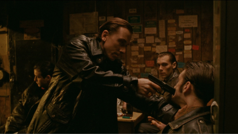 Cena de Fuga para Odessa. Plano médio tenso no interior de uma sala rústica com paredes de madeira cobertas de papéis e notas. Em destaque, Tim Roth, vestindo um casaco de cabedal preto, aponta com firmeza uma pistola preta diretamente à boca de um homem encurralado contra a parede. A vítima inclina a cabeça para trás em aflição, enquanto outros dois cúmplices ao fundo observam a cena com apreensão num ambiente escuro e com iluminação avermelhada.