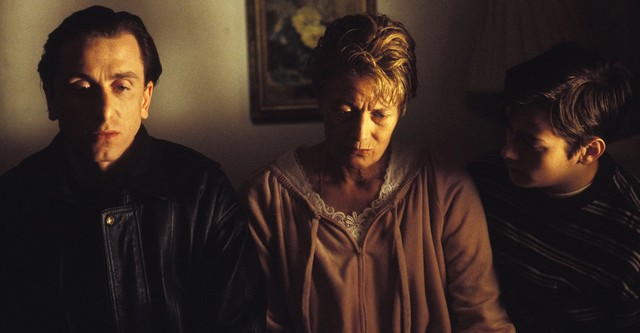 Cena de Fuga para Odessa. Plano médio de três personagens lado a lado num interior mal iluminado, banhados por uma luz frontal quente e suave. À esquerda, Tim Roth veste um blusão preto e olha em frente com um olhar sombrio e abatido. Ao centro, Vanessa Redgrave, mulher branca de cabelo curto loiro e casaco felpudo cor-de-rosa claro, mantém a cabeça ligeiramente inclinada para baixo com uma expressão de cansaço ou tristeza profunda. À direita, Edward Furlong olha fixamente para a mãe.