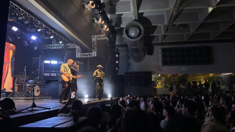 Registro do show de Os Garotin. À esquerda da imagem, com perspectiva da lateral do palco, o trio está na plataforma sob uma iluminação azul. Cupertino toca guitarra. À direita, os fãs assistem à apresentação. Alguns levantando o smartphone para tirar fotos e gravar vídeos.