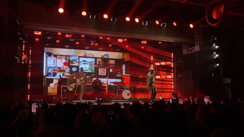Registro do show de Os Garotin. Sobre o palco estão o trio, o baterista Felipão e o baixista Júlio Raposo. O palco tem iluminação mais avermelhada, com uma imagem projetada no telão, que mostra um mural com elementos que fazem referência à estética do álbum Força da Juventude. É possível ver alguns celulares gravando o show.