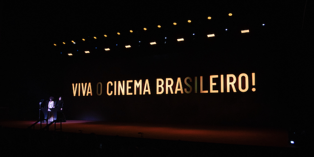 Uma imagem de um palco com quase todas as luzes apagadas e a frase “Viva o Cinema Brasileiro!” na tela em amarelo. No canto esquerdo é possível identificar pessoas no palco. As cores que prevalecem é preto e amarelo.