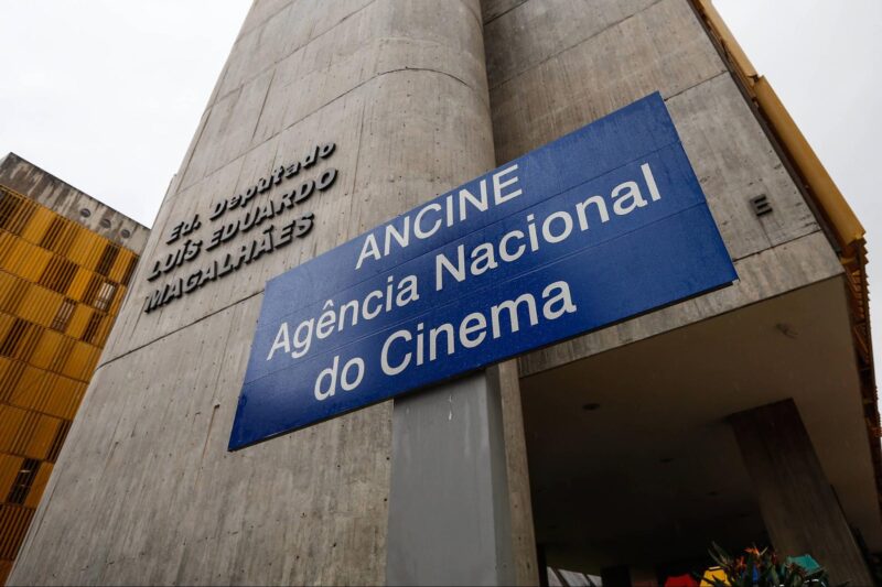Foto tirada do prédio da Agência Nacional do Cinema. Com o ângulo de baixo, ela foca na placa azul da agência, que contrasta com a parede cinza.