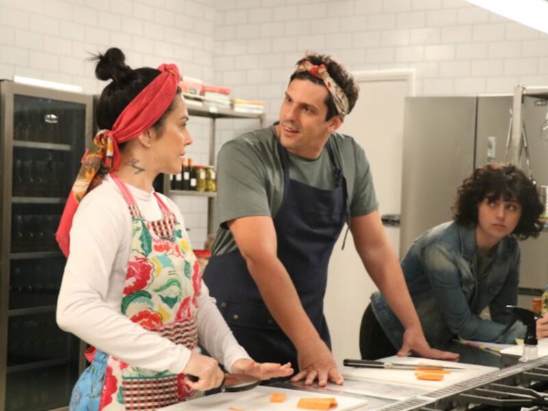 Três pessoas colaboram em uma cozinha profissional. Cleo Pires, à esquerda, com um lenço vermelho vibrante e avental floral, foca em fatiar cenouras. Ela troca olhares com Rafael Infante, que veste um avental azul e uma bandana, e está apoiado na bancada com uma expressão curiosa e engajada. Uma terceira pessoa, mais jovem e de jaqueta jeans, observa a interação em silêncio à direita. A cozinha é moderna, com equipamentos de inox e prateleiras organizadas ao fundo.