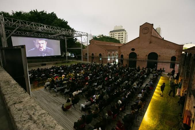 Imagem da Cinemateca em São Paulo. Contém uma tela projetada para um festival, com cadeiras dispostas à sua frente. Pessoas sentadas assistem ao filme que passa, com uma iluminação que indica o fim da tarde. É uma foto tirada de uma posição levemente distante da atividade.