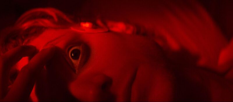 Cena do filme Cara-de-Barro Na imagem, o protagonista está deitado enquanto uma mão abre seus olhos que estão com a pupila dilatada. Apenas metade do seu rosto aparece no quadro e há uma iluminação vermelha em cena. 