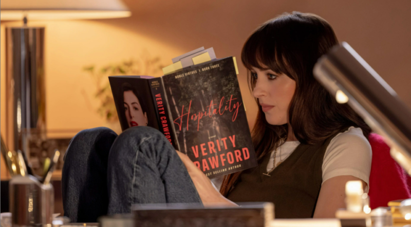 Uma mulher branca de cabelo castanho e franja está sentada com as pernas dobradas e os braços apoiados nela. Em suas mãos, há o livro “Hospitality” de Verity Crawford. Na contracapa, está destacada a foto da autora, uma mulher de cabelo escuro e batom vermelho. O fundo é iluminado por uma luminária com luz alaranjada e o primeiro plano exibe uma mesa com objetos desfocados.