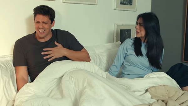 Uma cena de quarto com duas pessoas na cama. À esquerda, Infante com cabelo escuro e uma camiseta cinza está sentado com uma expressão de dor visível, segurando o peito com a mão direita e olhando para a frente com angústia. À direita, Cleo com longo cabelo preto e uma blusa azul-clara de mangas compridas está sentada, olhando para o homem com uma expressão de preocupação e confusão, os lábios entreabertos. Eles estão em uma cama de casal com lençóis brancos e travesseiros brancos, e uma coberta bege está visível nos pés da cama. A parede branca atrás deles tem três molduras simples em tons de madeira clara e branco penduradas. O ambiente é bem iluminado, sugerindo luz natural suave.