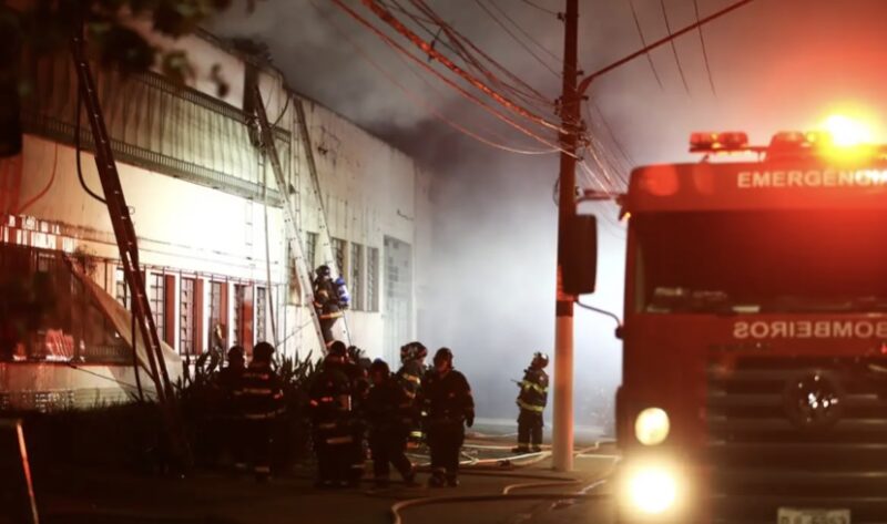 Imagem do último incêndio ocorrido na Cinemateca em São Paulo (2021). A foto é dividida entre o prédio em chamas e o veículo de bombeiros, com profissionais entrando no local. A iluminação é escura, com bastante fumaça e luzes vermelhas que sinalizam a presença do corpo de bombeiros.