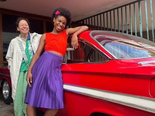 Liliane à esquerda, de pele clara e cabelos escuros curtos, está de pé atrás do carro e ri com a boca aberta. Ela veste uma jaqueta off-white sobre uma camisa verde estampada, calça verde folgada e uma alça de bolsa em preto e branco. Barbara Maria à direita, de pele negra e cabelo afro escuro com uma flor rosa presa, está apoiada no teto do carro com a mão direita e sorri abertamente. Ela usa um cropped laranja e uma saia roxa plissada. O carro está em uma área externa, ao lado de um prédio com janelas grandes e uma cerca preta ao fundo. A luz do dia brilha nas laterais do carro.