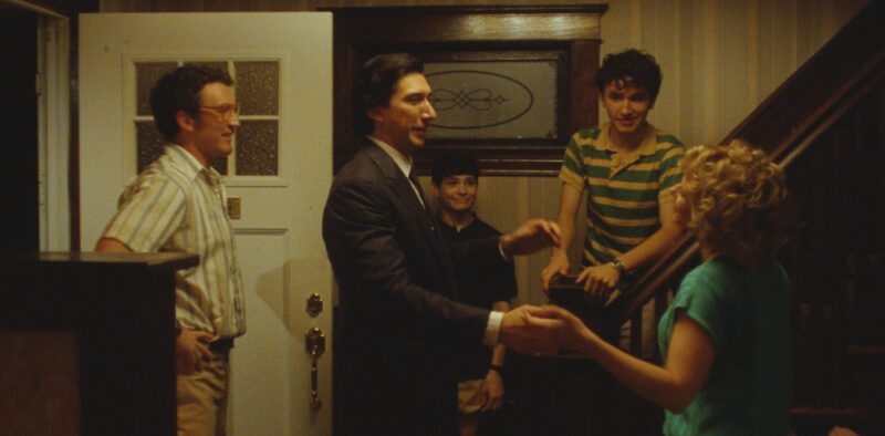 Cena do filme Paper Tiger.Plano médio em tom amarelado. Ao centro, Adam Driver, homem branco de terno escuro e cabelo castanho escuro, segura as mãos de Scarlett Johansson, uma mulher branca que está de costas no canto inferior direito, que possui cabelo loiro cacheado e veste uma camisa azul-esverdeada. À esquerda, em pé perto de uma porta branca, Miles Teller, um homem branco de óculos e camisa listrada de mangas curtas, sorri. Ao fundo e à direita, na escada de madeira da casa, dois jovens sorriem observando a cena: um garoto branco de camisa polo azul-marinho e outro mais alto usando camisa polo listrada de verde e amarelo debruçado no corrimão. 
