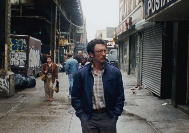 Cena do filme Paper Tiger. Plano geral granulado e cinematográfico no nível da rua. O homem com jaqueta bomber azul e óculos caminha de costas por uma rua urbana completamente invadida por uma densa vegetação selvagem, que cobriu o asfalto quebrado e os pilares de metal do trem elevado acima. O caminhão à esquerda está quase invisível, e as lojas à direita estão corroídas e cobertas por plantas.