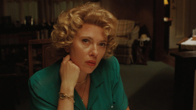 Cena de Paper Tiger. Plano médio de Scarlett Johansson, mulher branca com cabelo loiro cacheado estilo vintage. Ela olha seriamente para a câmera. Ela veste uma blusa verde-escura e tem a mão apoiada no rosto, usando um relógio de ouro no pulso e um colar fino. O fundo é uma sala de estar com móveis de madeira e um sofá xadrez sob iluminação ambiente quente. 