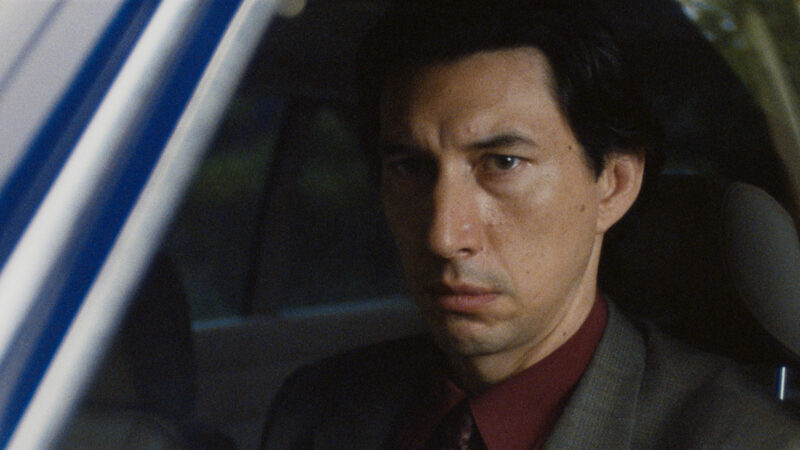 Cena de Paper Tiger: O personagem Gary está no interior de um veículo. Ele está sentado no banco do motorista, vestindo terno cinza, camisa social vinho e gravata escura. Seu olhar está fixo para a frente com uma expressão tensa, séria e pensativa. À esquerda do enquadramento, vê-se a coluna do carro e a janela, através da qual se nota uma iluminação externa suave e azulada, indicando ser final de tarde ou início da noite. A luz interior é fraca e sutil, destacando as feições marcantes e os olhos do ator.