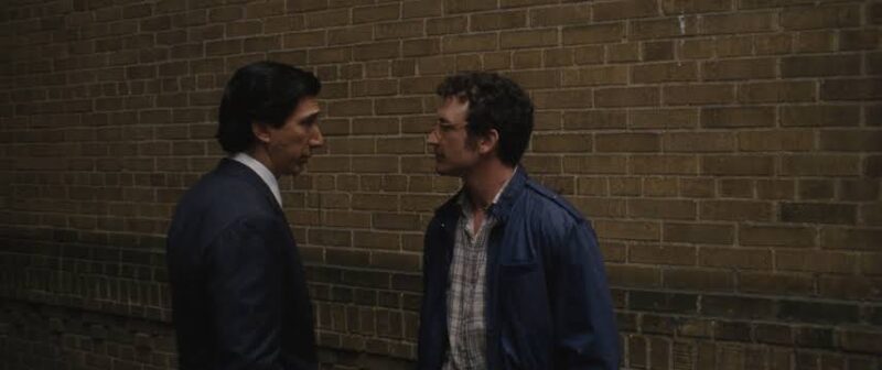 Cena de Paper Tiger.Plano médio de dois homens de perfil, um de frente para o outro, conversando em um beco junto a uma parede de tijolos marrons. À esquerda, Adam Driver veste um terno escuro sobre camisa branca, observa o outro homem com uma expressão séria e atenta. À direita, Miles Teller, com cabelo curto encaracolado e jaqueta azul-escura sobre uma camisa de xadrez claro, olha diretamente para Adam Driver enquanto fala. A iluminação do local é suave.