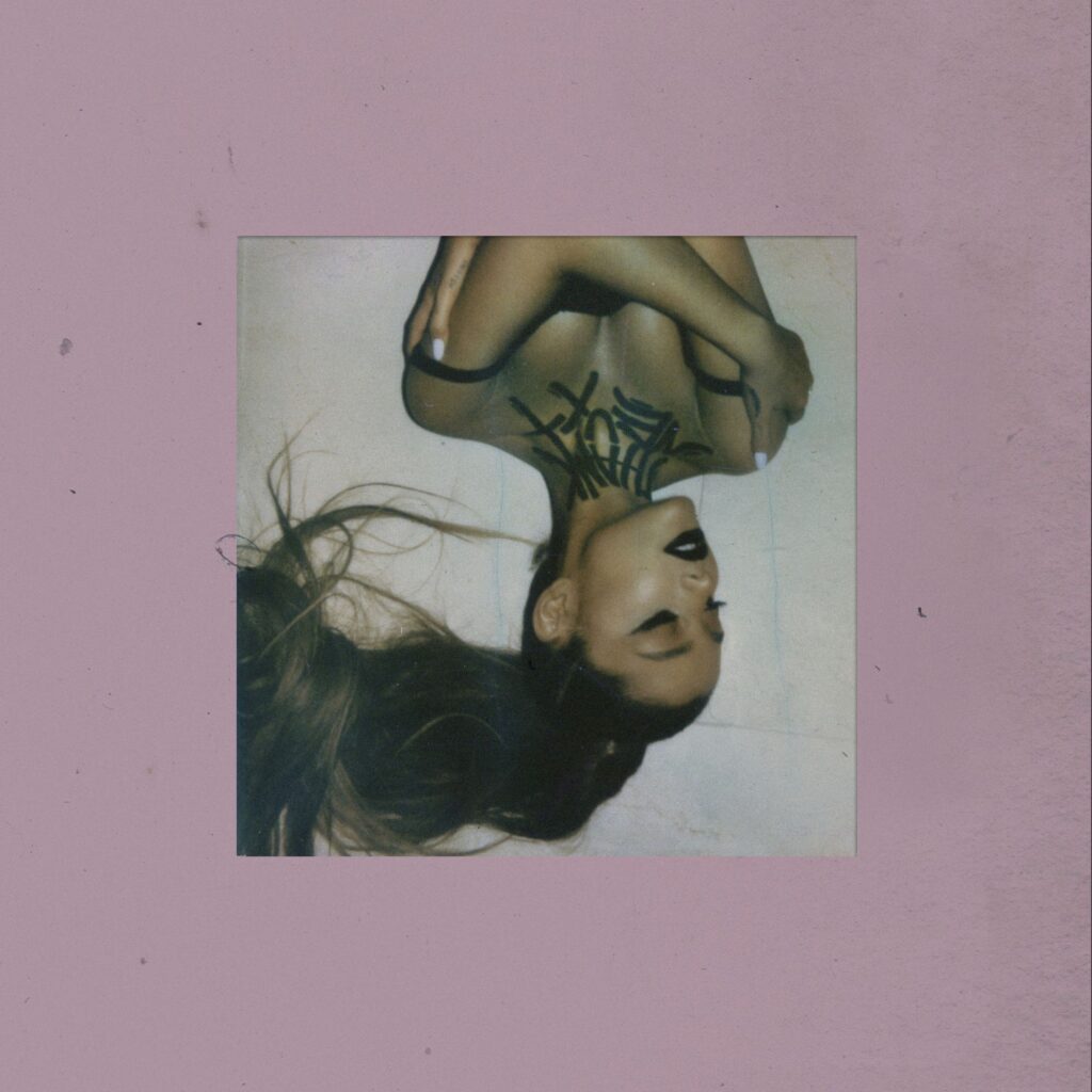 Capa do álbum "thank u, next" da cantora Ariana Grande. Ela está deitada de costas, parcialmente de cabeça para baixo na imagem. Ariana está com os olhos fechados e um sorriso suave nos lábios. Ela usa um batom escuro, um delineado preto e seu cabelo comprido está solto, caindo para baixo. No pescoço e no peito de Ariana, há escritas em preto, formando o nome do álbum. O fundo da imagem é de uma cor pastel suave, um tom de rosa claro.
